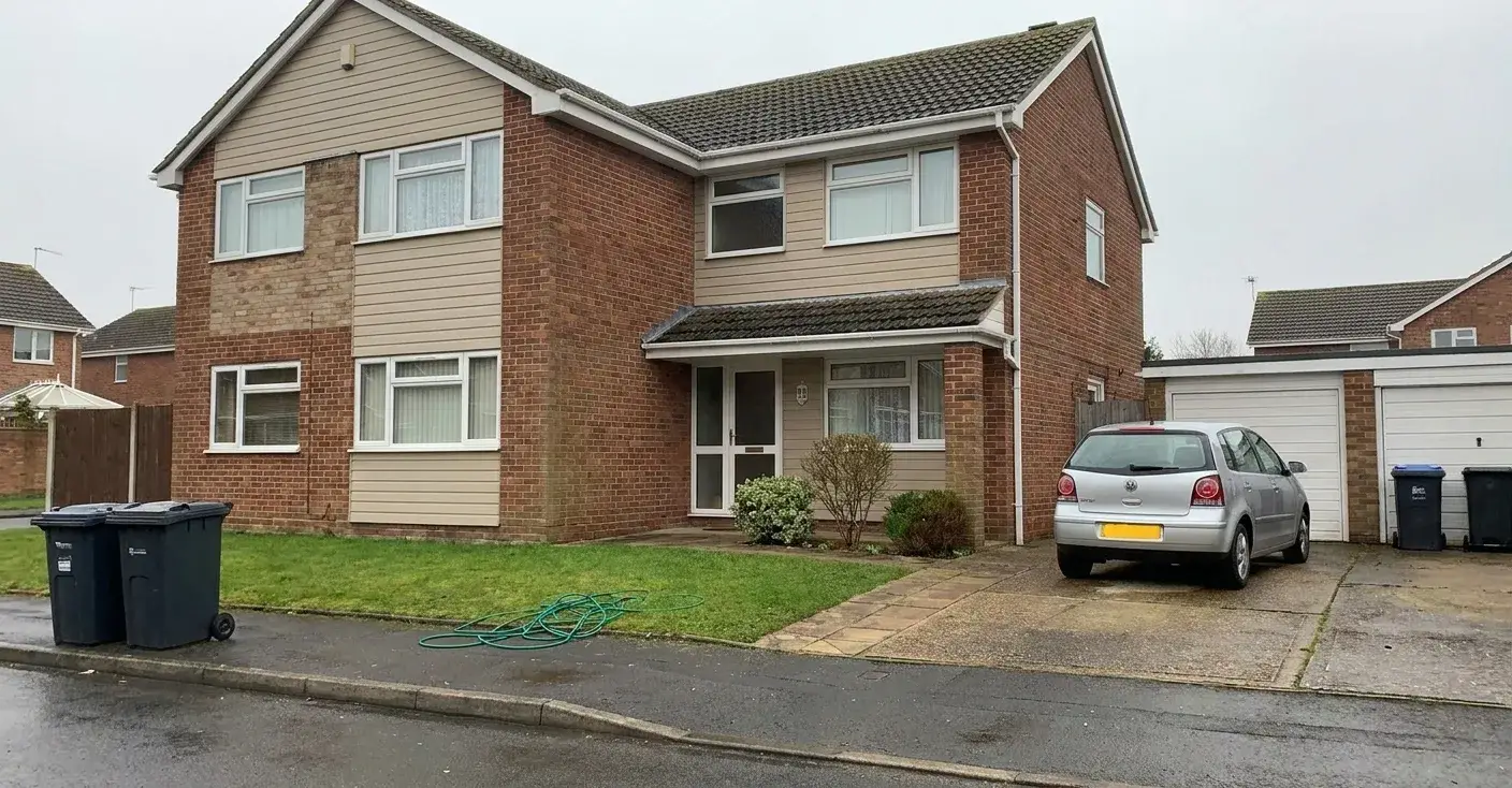 A suburban house photographed under a flat grey sky, with a car on the driveway, bins at the kerb and a hose on the lawn
