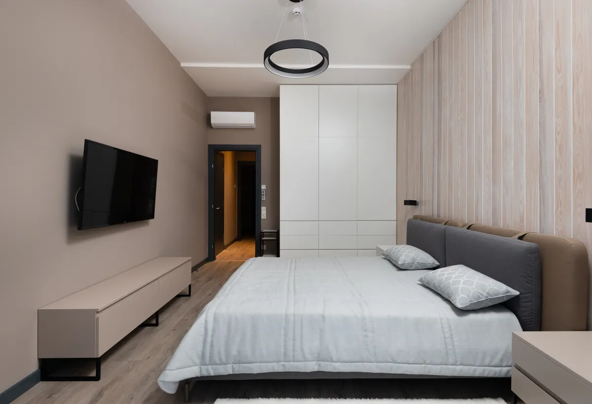 A staged bedroom with a wide bed, an upholstered headboard, a wall-mounted TV and a low media console