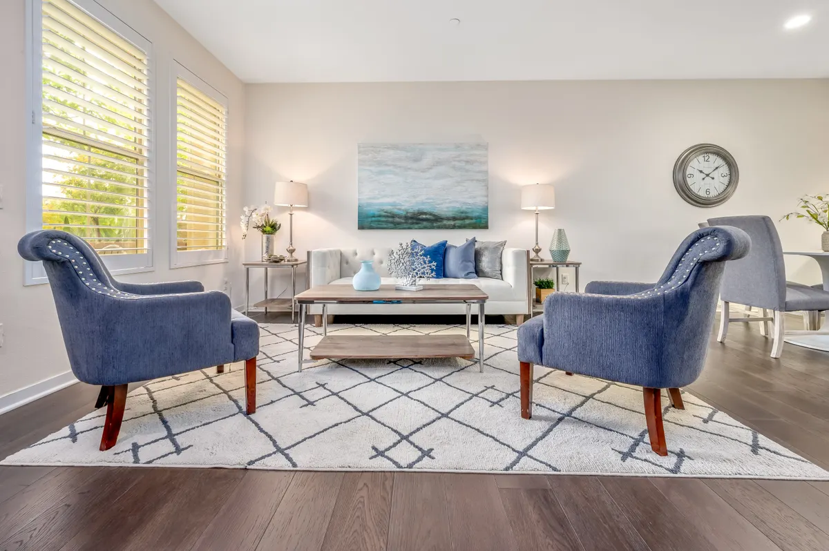 A staged living room with two blue armchairs facing a white tufted sofa, a patterned rug and coastal artwork