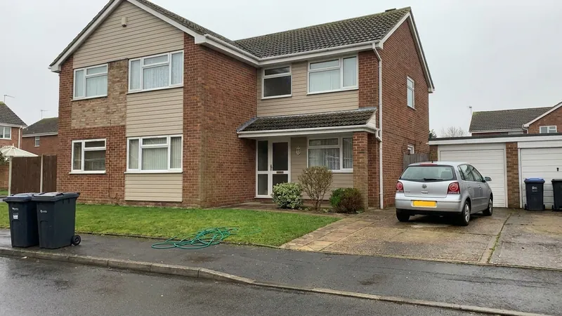 A house under a flat grey sky with a silver car on the driveway, two wheelie bins at the kerb and a green hose coiled on the lawn